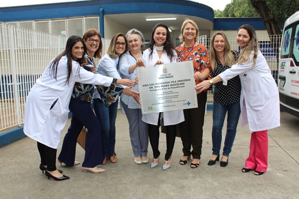 #PraCegoVer: Parte da equipe da UBS Integrada Vila Oratório, oito mulheres, segura a placa da entrega da reforma na fachada da unidade